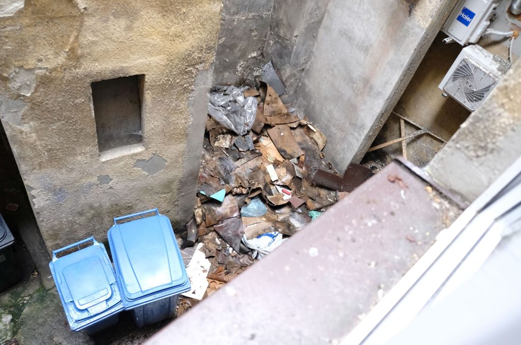 Widok z gór na podwórko wewnątrz kamienicy, dwa niebieskie pojemniki na śmieci obok walające się stosy, złożonych, zawilgoconych, szarobroązowych kartonów po opakowaniach 