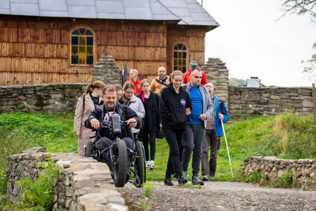 Grupa turystów z niepełnosprawnym na wózku elektrycznym na tle drewnianego kościoła