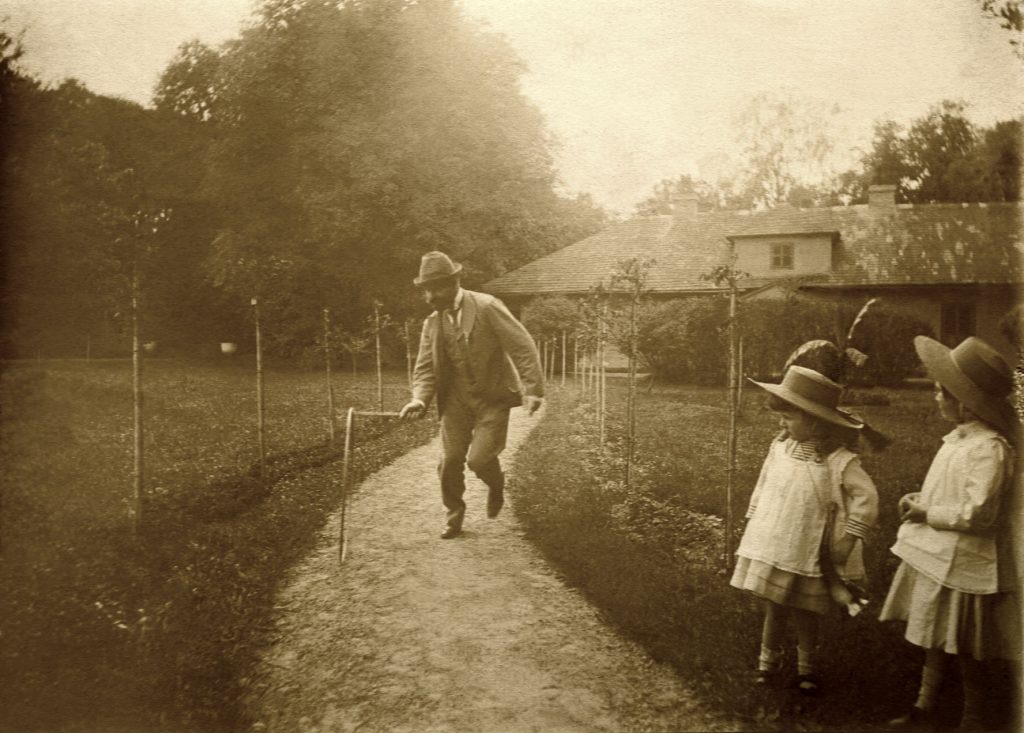 Na zdjęciu znajduje się mężczyzna. który toczy koło, obok przy ścieżce stoją dwie dziewczynki w kapeluszach.